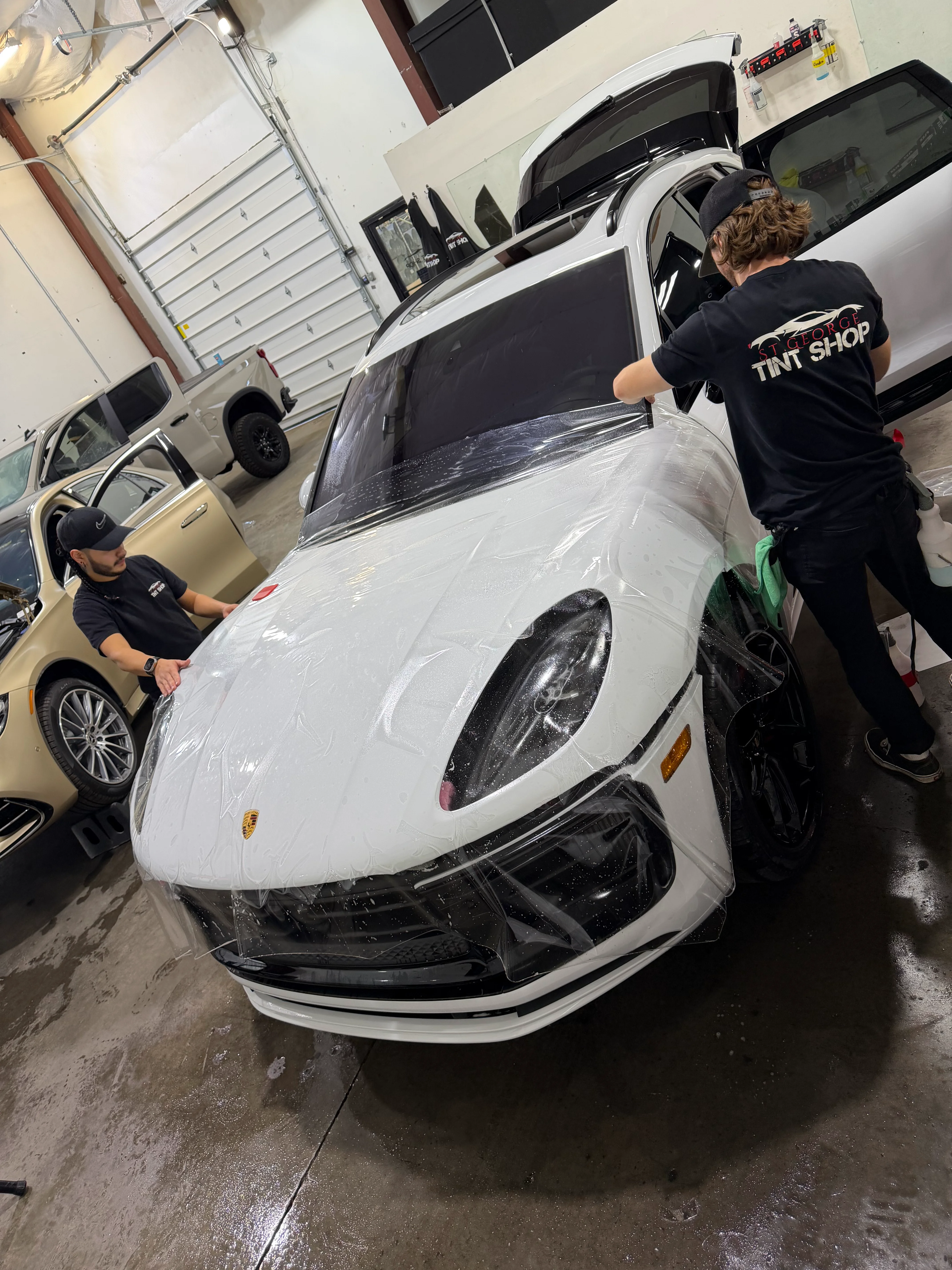 White Porsche Macan SUV receiving paint protection film (PPF) application by skilled technicians at a tint shop in St. George, Utah.